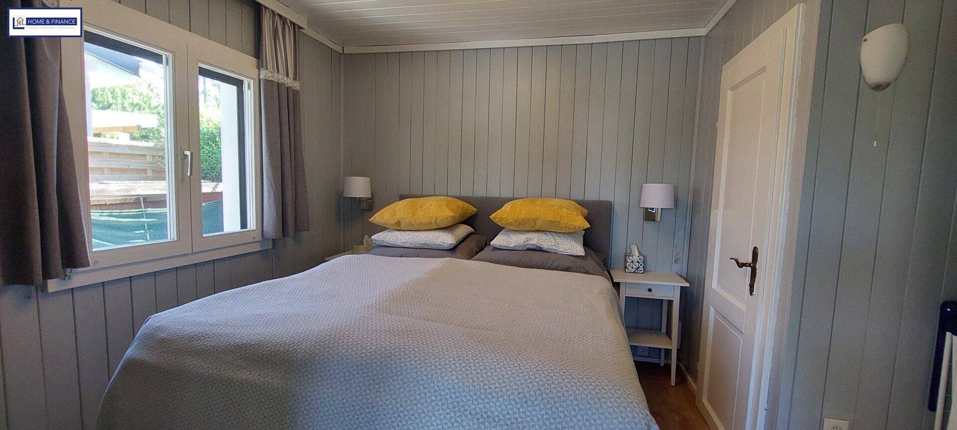 Helles Schlafzimmer mit Doppelbett, grauen Holzpaneelen an den Wänden und einem Fenster.