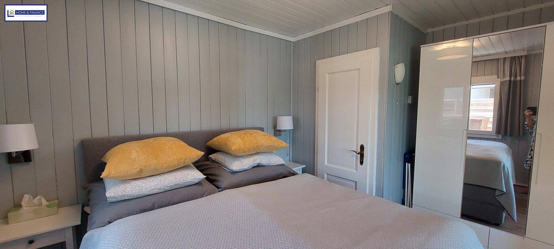 Schlafzimmer mit Doppelbett, grauen Paneelwänden und einem weißen Kleiderschrank mit Spiegeltür.