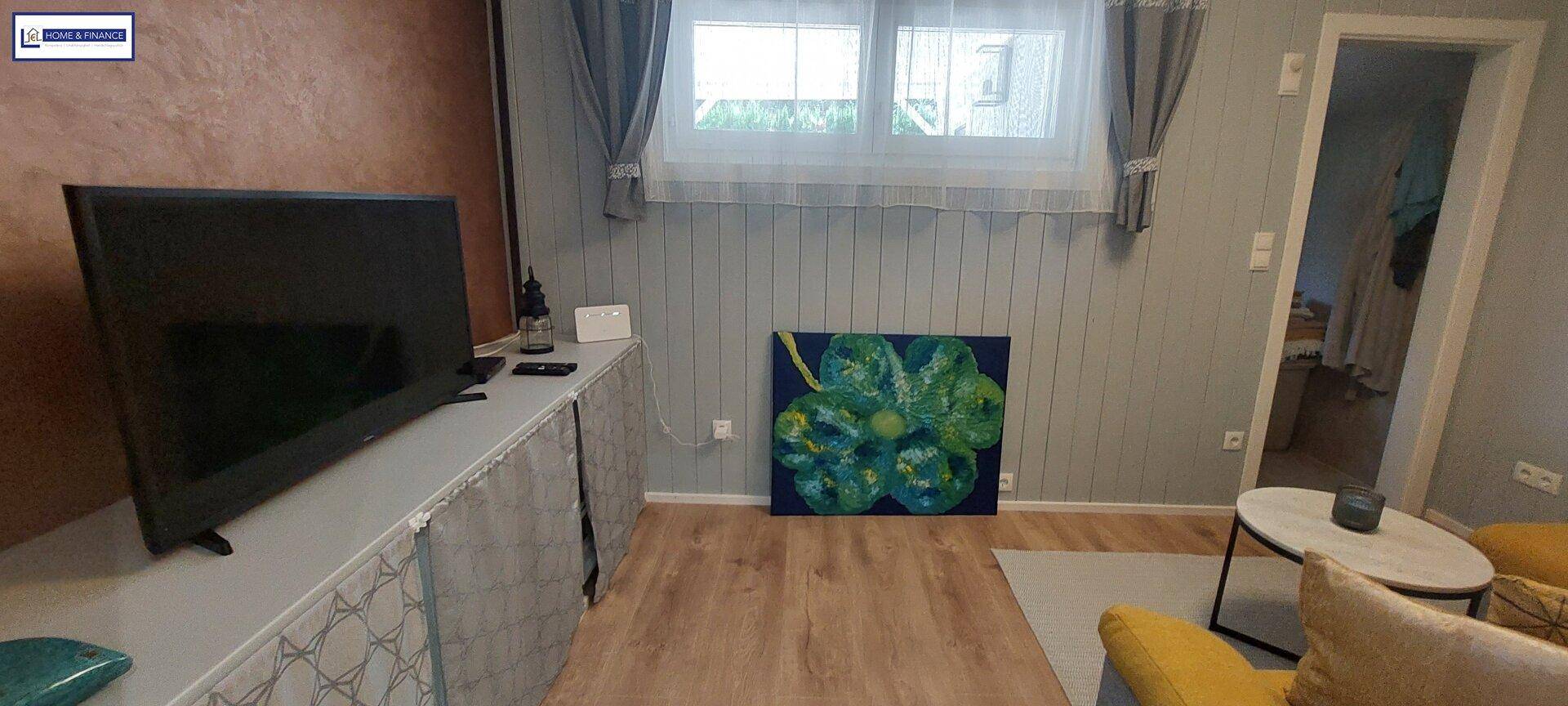 Gemütliches Wohnzimmer mit Fernseher, Holzfußboden und Fenster mit Blick nach draußen.