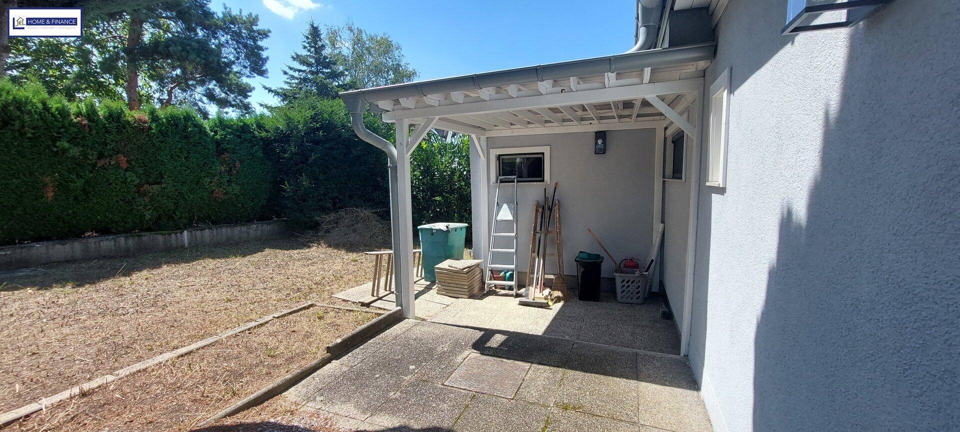 Überdachte Terrasse mit Werkzeugen und Leiter neben dem Haus, umgeben von trockenem Rasen.