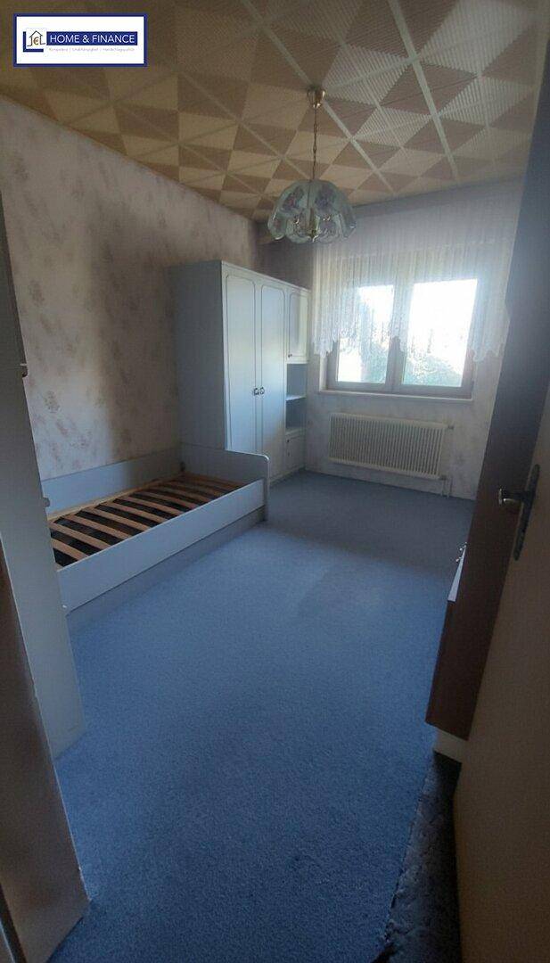 Schlafzimmer mit blauem Teppichboden, Fenster und einem einfachen Bettgestell.