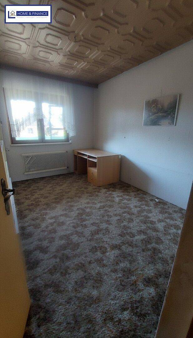 Schlafzimmer mit gemustertem Teppichboden, Schreibtisch und Fenster mit Tageslicht.