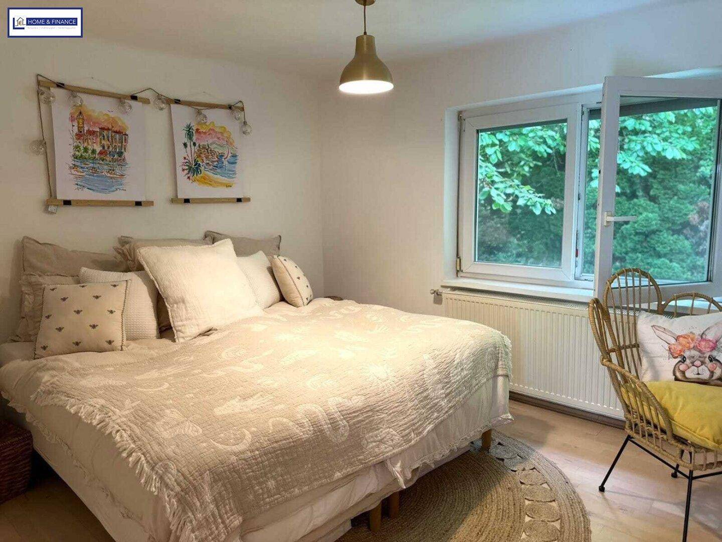 Helles Schlafzimmer mit großem Bett, Fenster mit Blick ins Grüne und Rattanmöbeln.