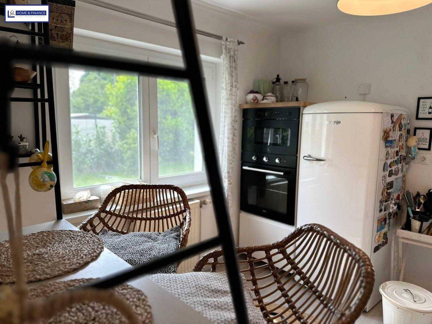 Heller Essbereich mit Rattan-Stühlen, Blick ins Grüne und integriertem Backofen sowie Kühlschrank.