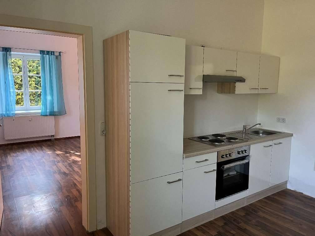 Kompakte Einbauküche mit weißen Fronten, Herd und Ofen, angrenzend an ein helles Zimmer mit Fenster und Holzboden.