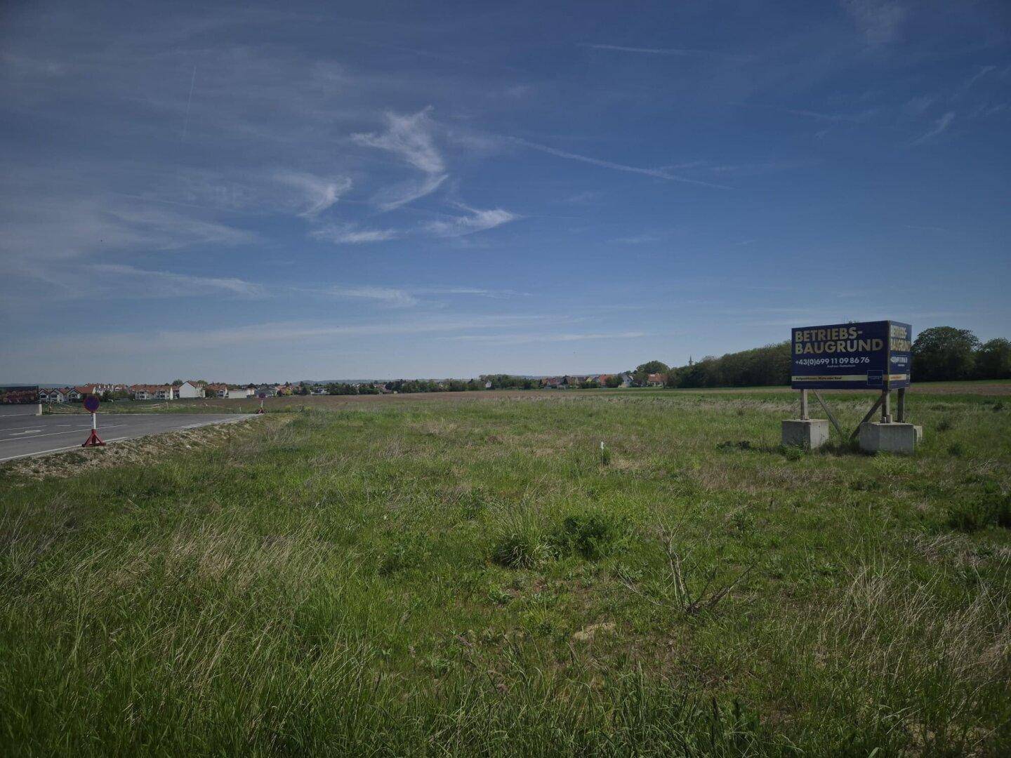 Großes unbebautes Feld mit einem Schild für Betriebs-Baugrund neben einer Straße unter klarem Himmel.