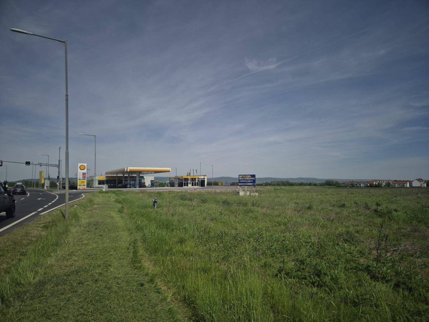 Weitläufiges unbebautes Grundstück neben einer Straße mit einer Tankstelle und einem Hinweisschild in der Ferne.