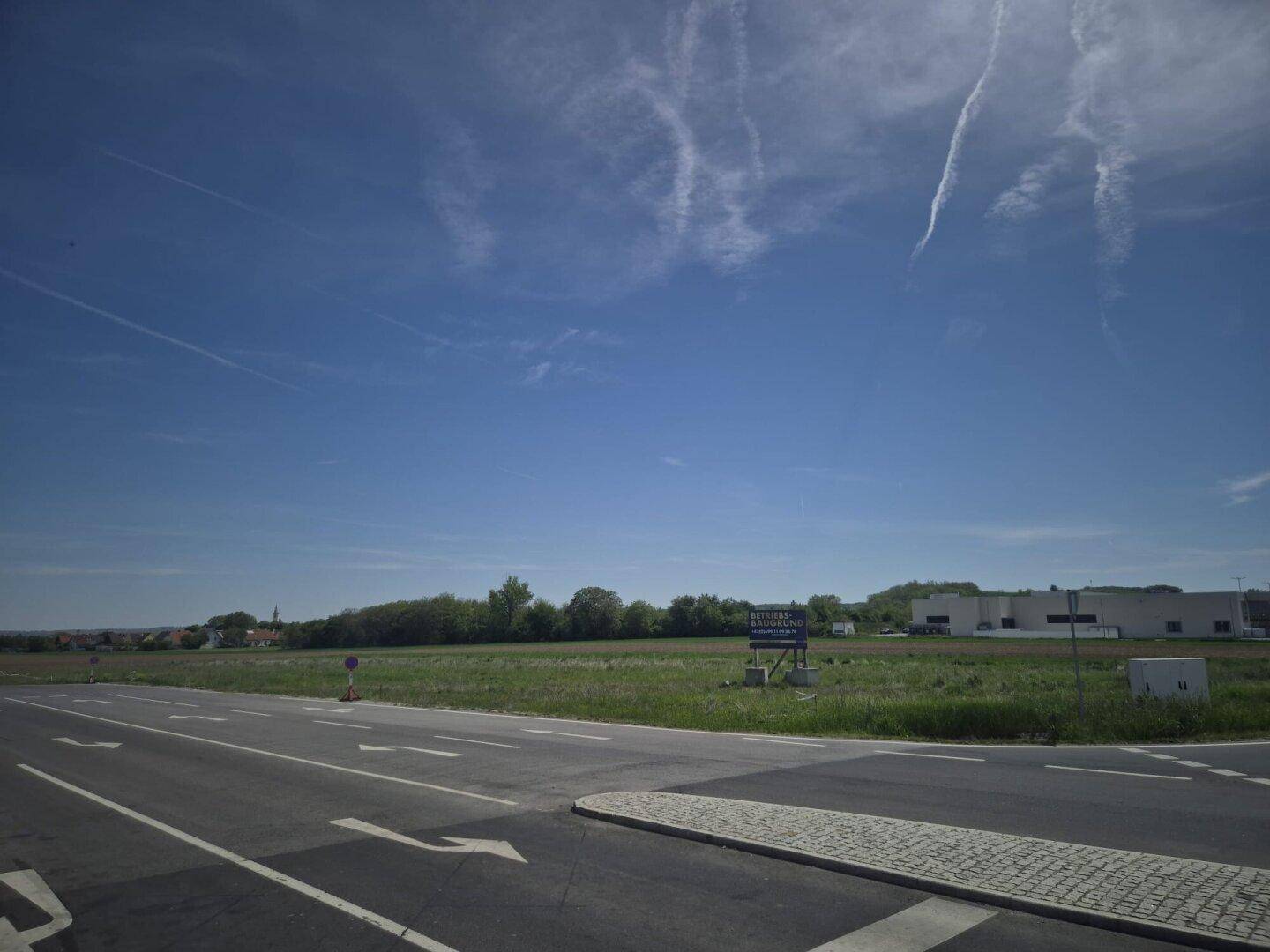 Unbebautes Feld an einer Straßenkreuzung mit Industriegebäuden und einem Hinweisschild in der Ferne.