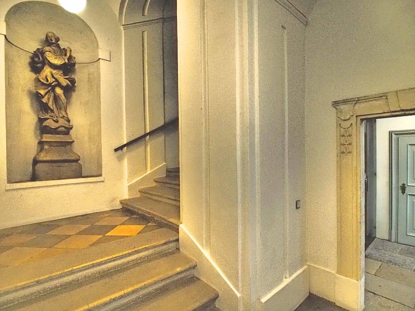 Historisches Treppenhaus mit einer Statue in einer Nische, gefliestem Boden und Holzgeländer.