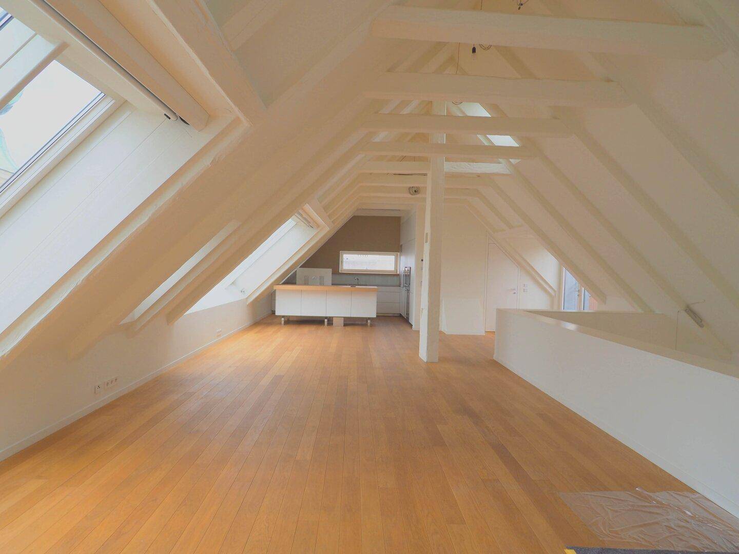 Heller Dachbodenraum mit sichtbaren Holzbalken, Holzboden und Dachfenstern, im Hintergrund eine Küchenzeile.