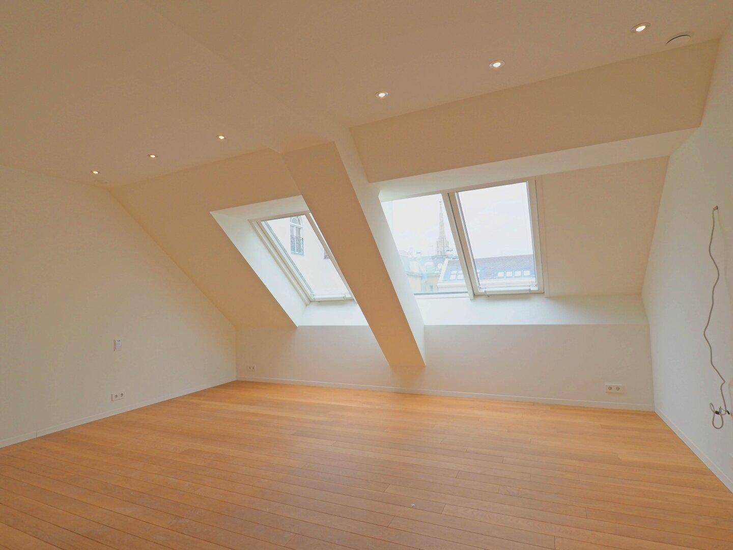 Geräumiges Zimmer im Dachgeschoss mit Holzboden, weißen Wänden und mehreren Dachfenstern.