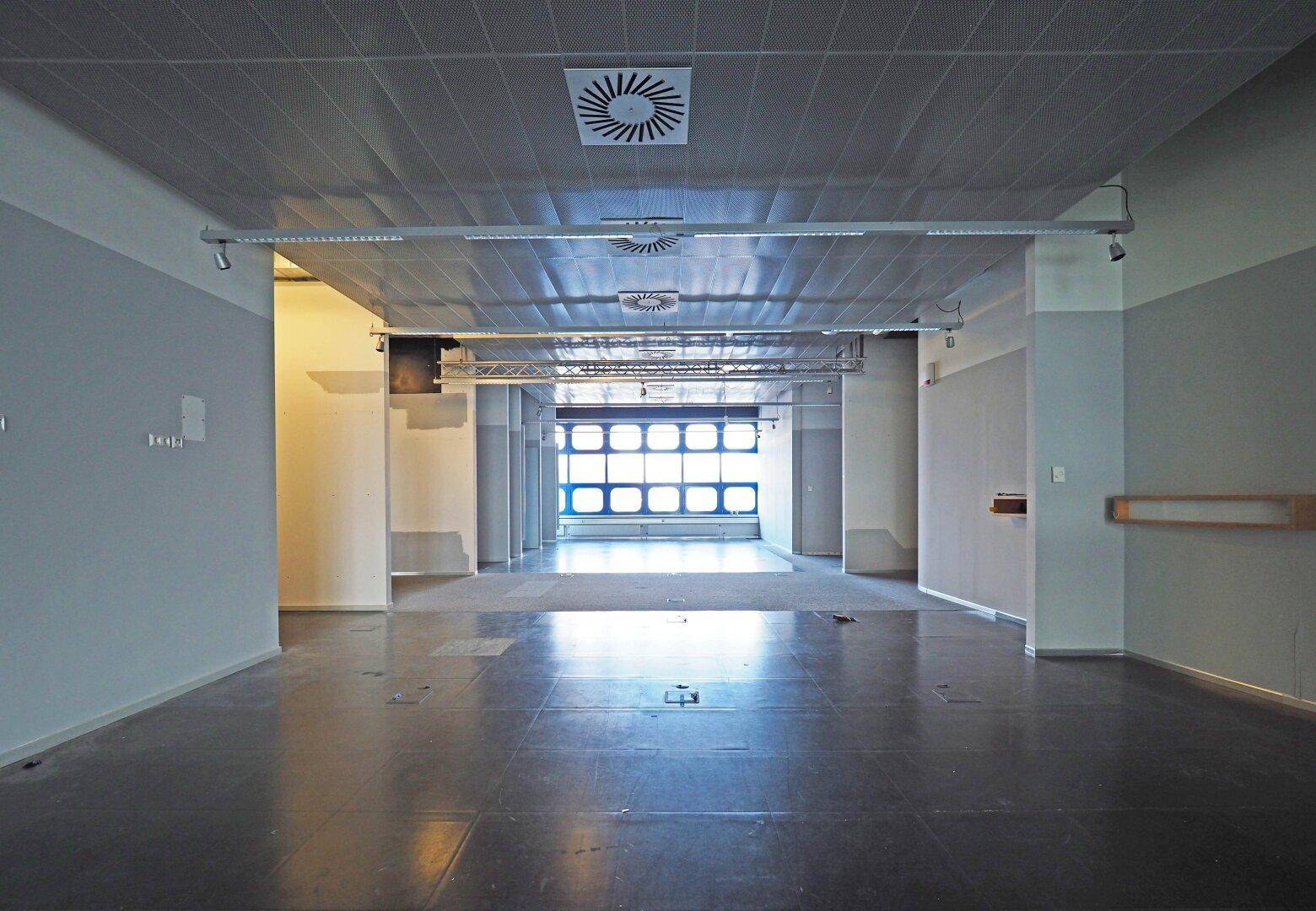 Leerer Gewerberaum mit abgehängter Decke, Lüftungsauslässen und Blick auf die Fensterfront.