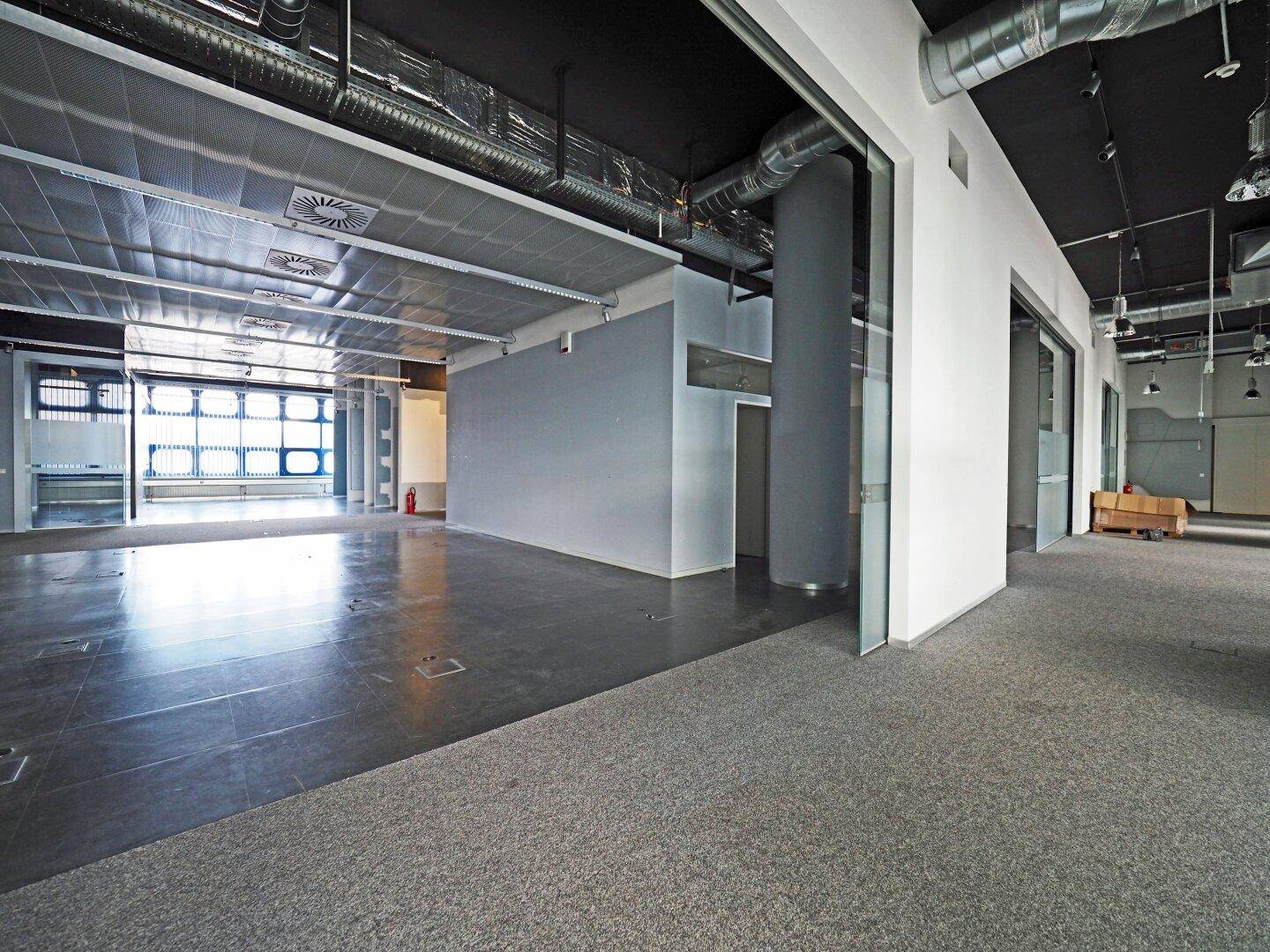 Geräumiger, leerer Bürobereich mit Teppich- und Fliesenboden, hohen Decken und sichtbaren Lüftungsrohren.
