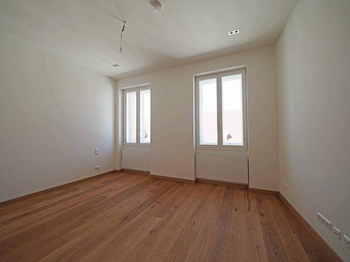 Leeres Zimmer mit zwei großen Fenstern und neu verlegtem Parkettboden, ideal als Schlafzimmer.
