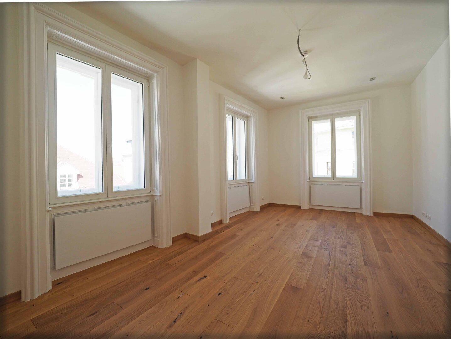 Helles Wohnzimmer mit drei Fenstern und elegantem Parkettboden, bereit zur individuellen Gestaltung.