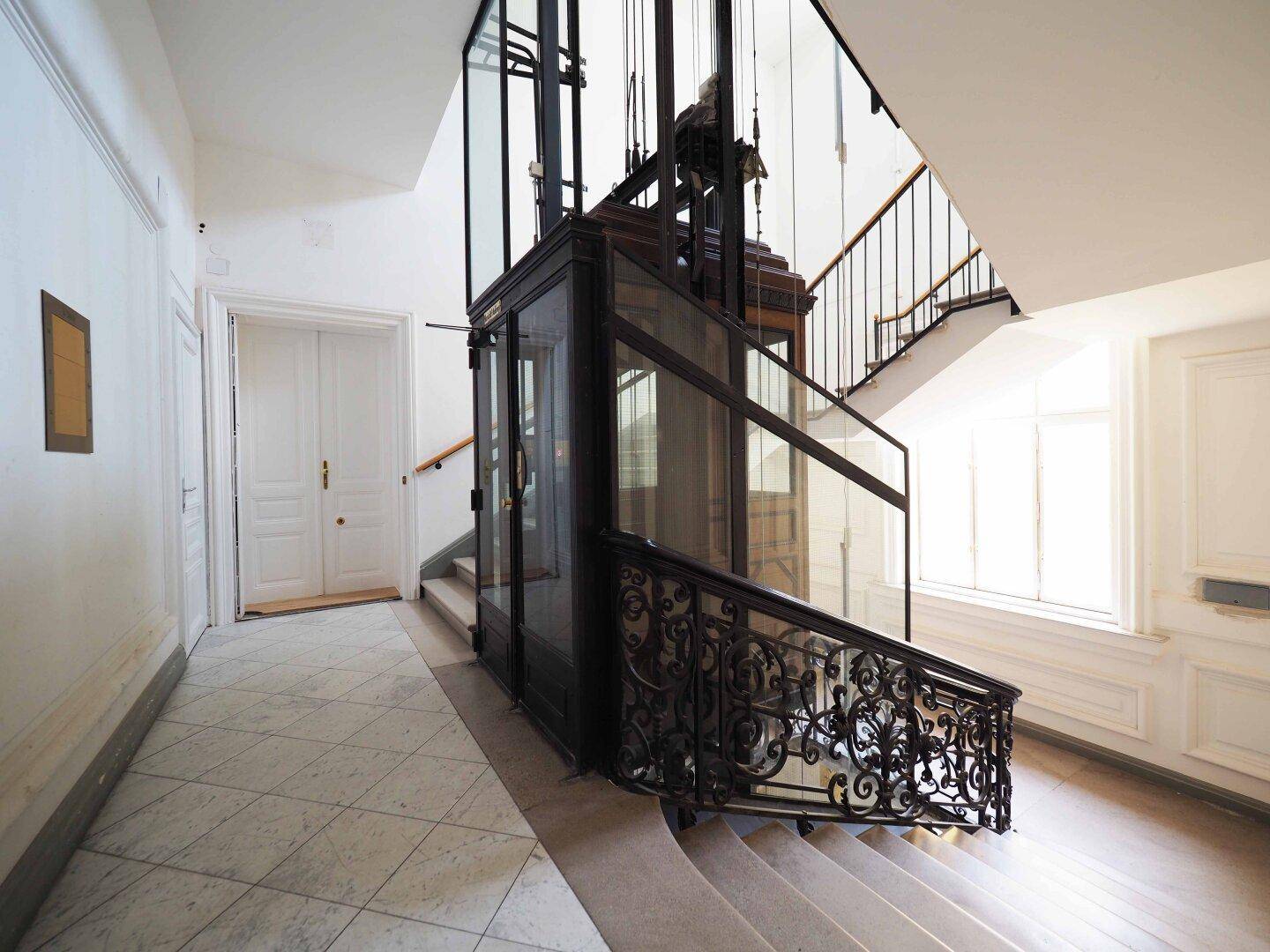 Historisches Treppenhaus mit kunstvollem Geländer und einem eleganten Glasaufzug.