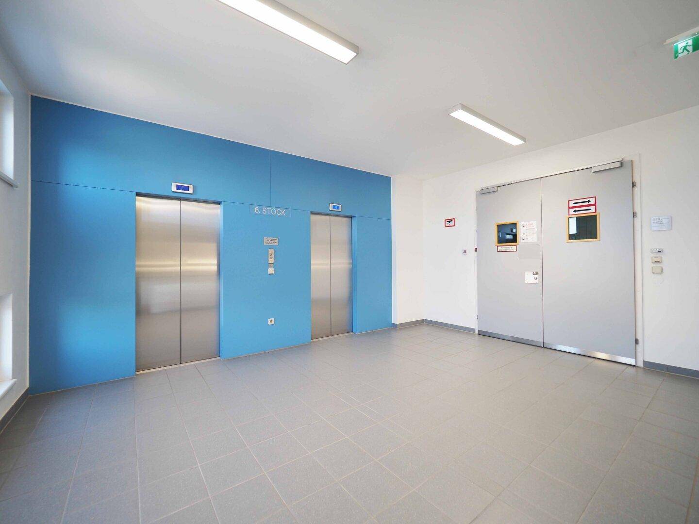 Moderner Aufzugsbereich mit zwei Edelstahlliften, blauer Akzentwand und hellen Fliesen auf dem Boden.