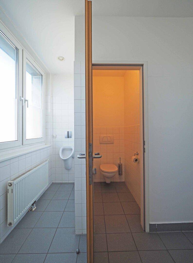Badezimmer mit Urinal, Fenster und einer separaten Toilettenkabine, alles in hellen Fliesen gehalten.