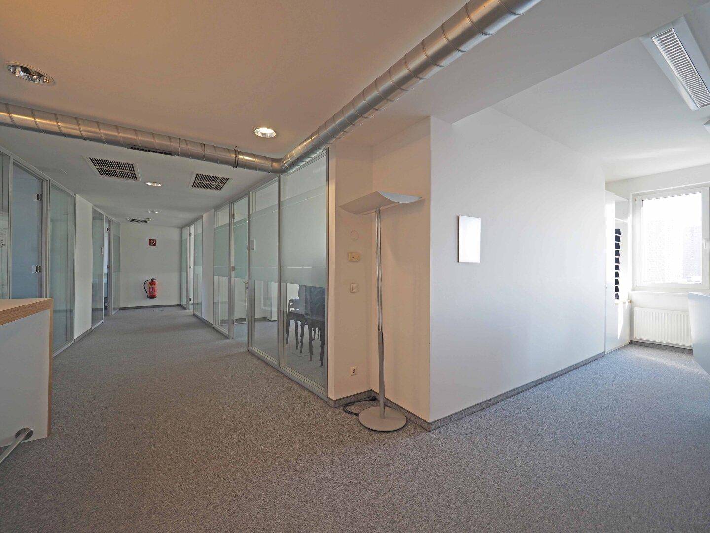 Eckbereich eines Bürogangs mit Teppichboden, Glaswänden und sichtbaren Lüftungsrohren an der Decke.