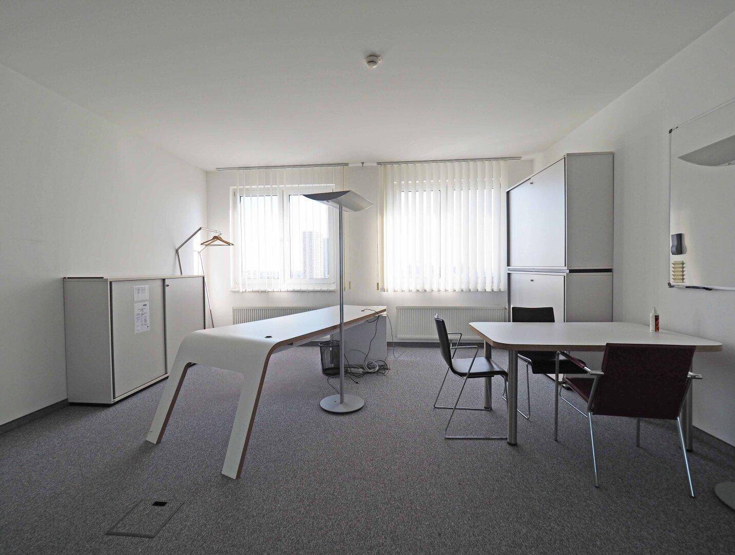 Helles Büro mit Teppichboden, modernen Schreibtischen und großen Fenstern mit Jalousien.