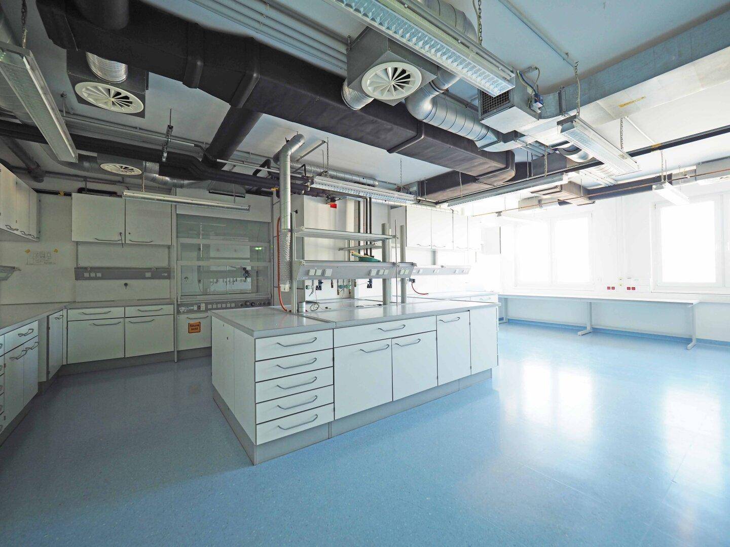 Großes Labor mit zentraler Insel, weißen Schränken, blauem Boden und sichtbarer Lüftungsanlage an der Decke.