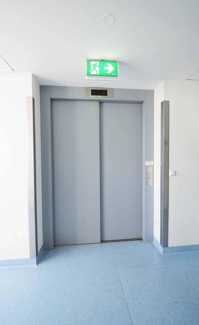 Nahaufnahme einer Aufzugstür mit Notausgangsschild darüber und blauem Bodenbelag.