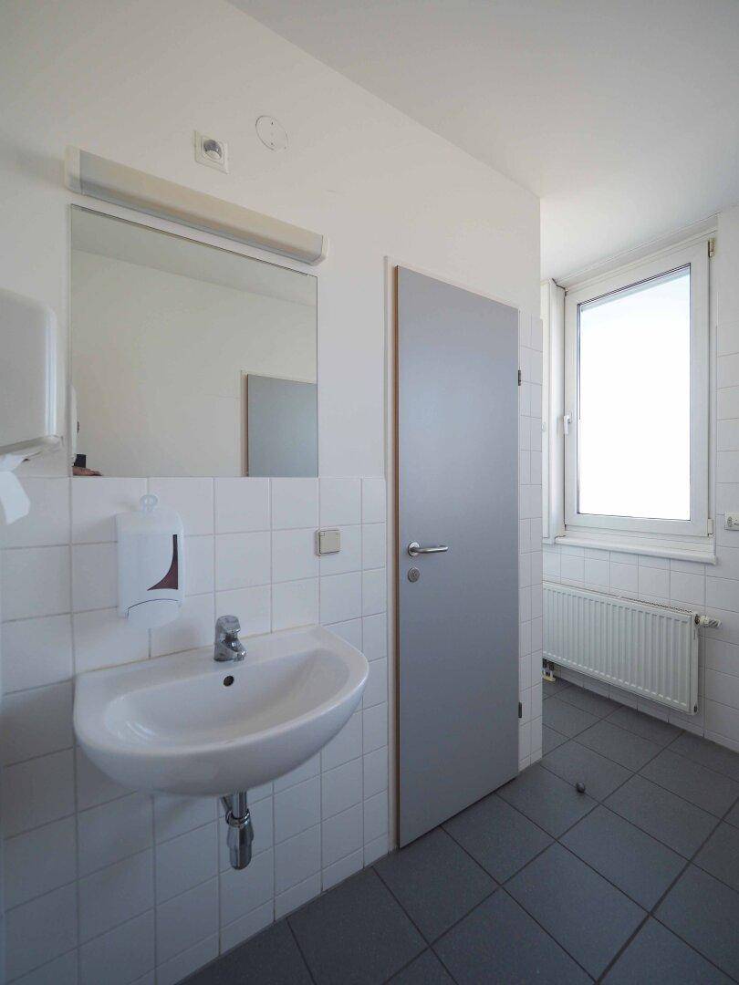Badezimmer mit Waschbecken, Spiegel, weißen Fliesenwänden, dunklem Fliesenboden und einem Fenster.