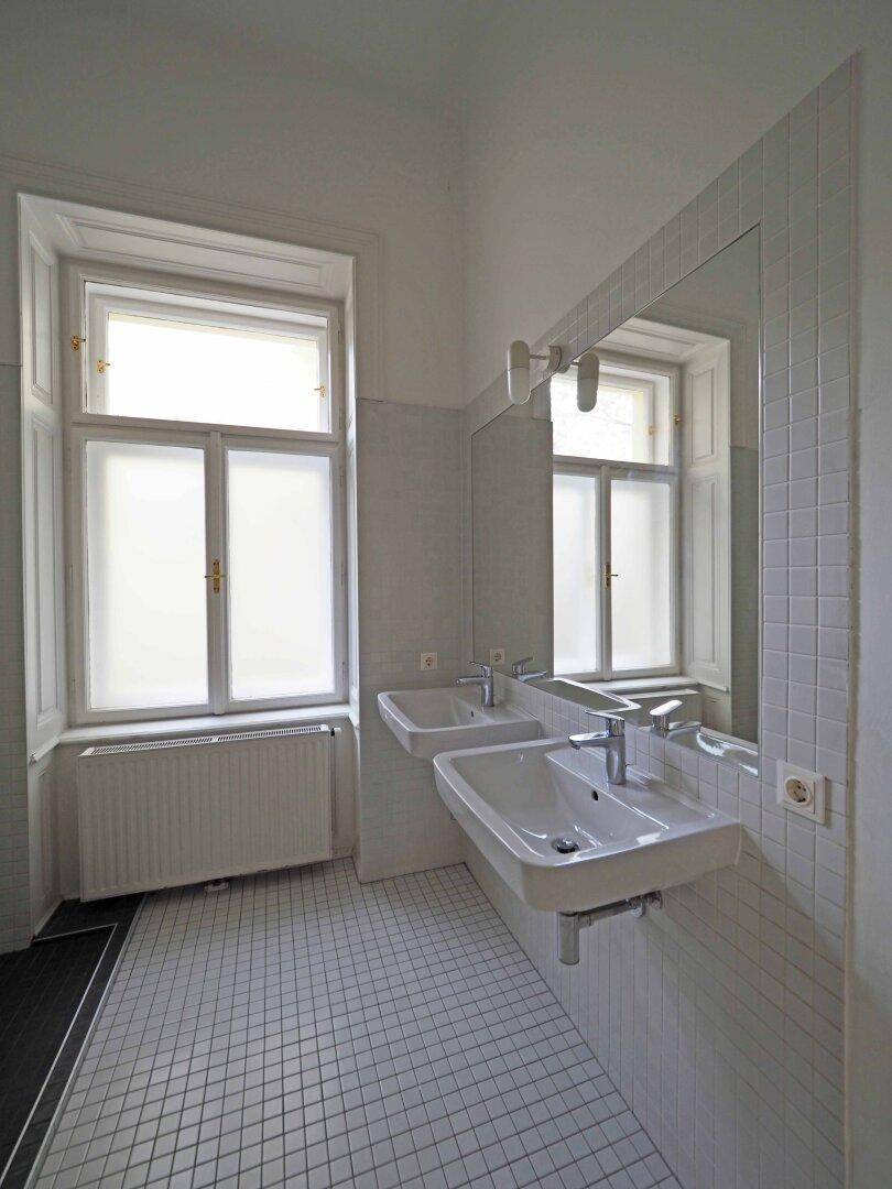 Helles Badezimmer mit zwei Waschbecken, weißen Fliesen und einem Fenster mit Milchglas.
