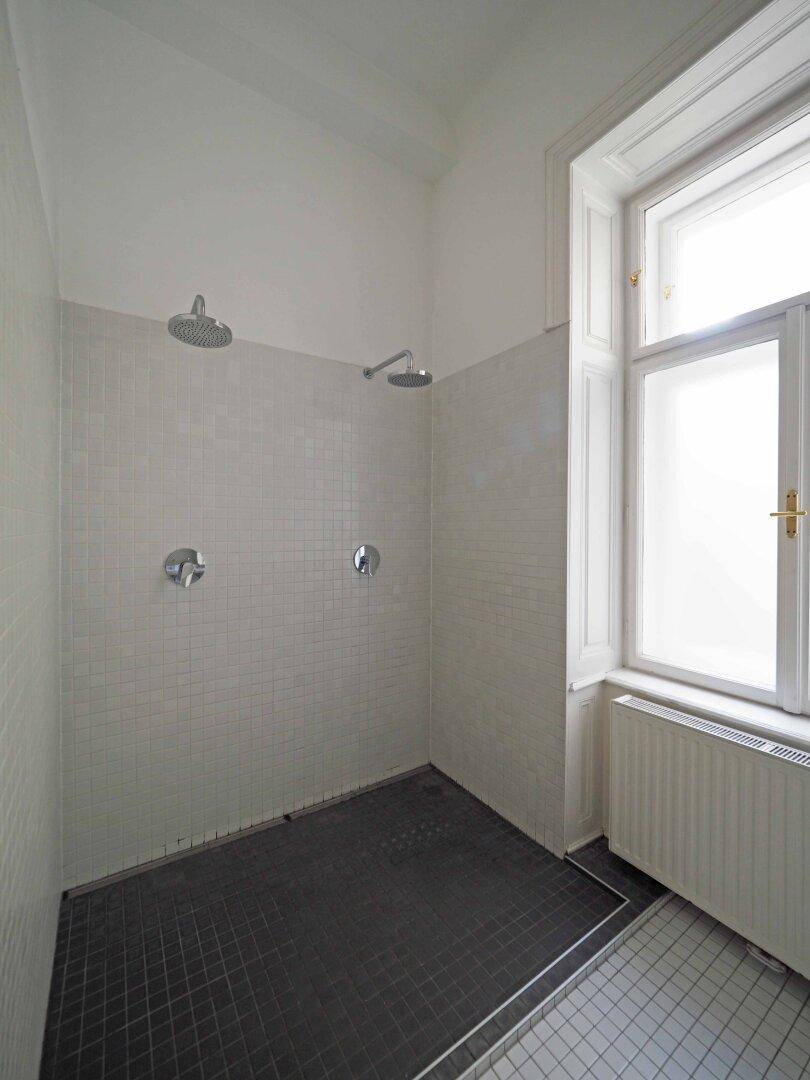 Duschbereich mit zwei Regenduschköpfen, weißen Fliesen und einem hohen Fenster.