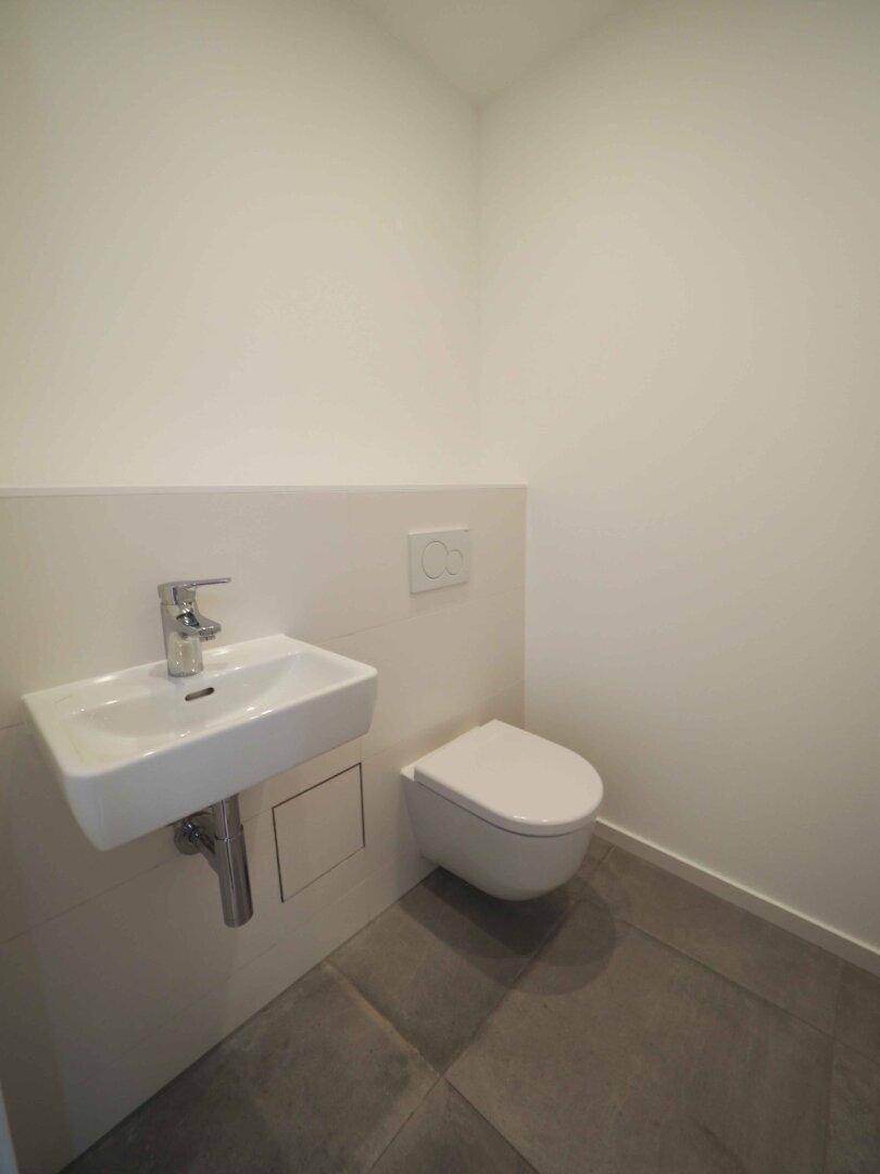 Modernes Gäste-WC mit wandhängender Toilette, kleinem Waschbecken und grauen Fliesen.