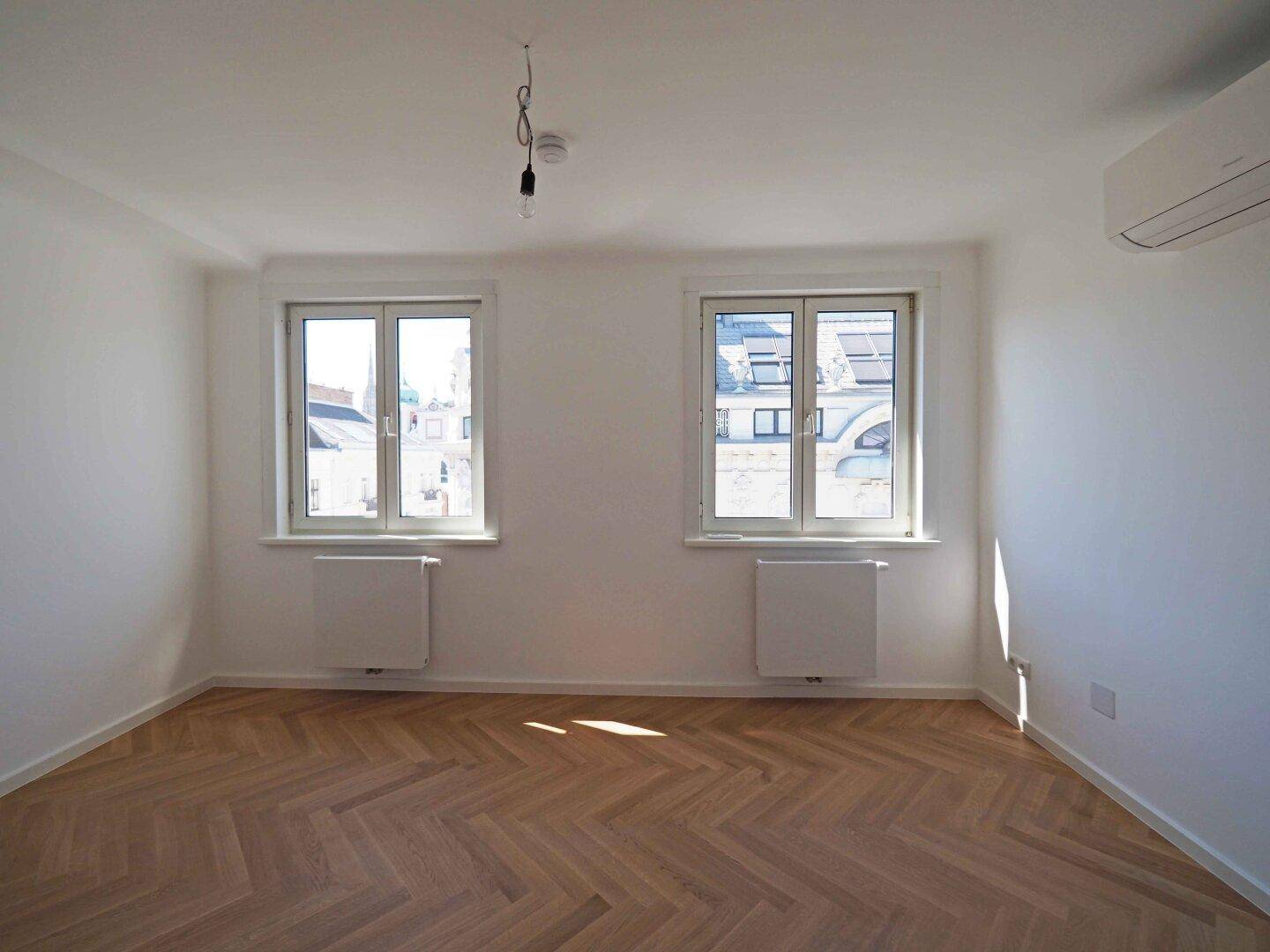 Geräumiges Zimmer mit elegantem Fischgrätparkett und zwei großen Fenstern.