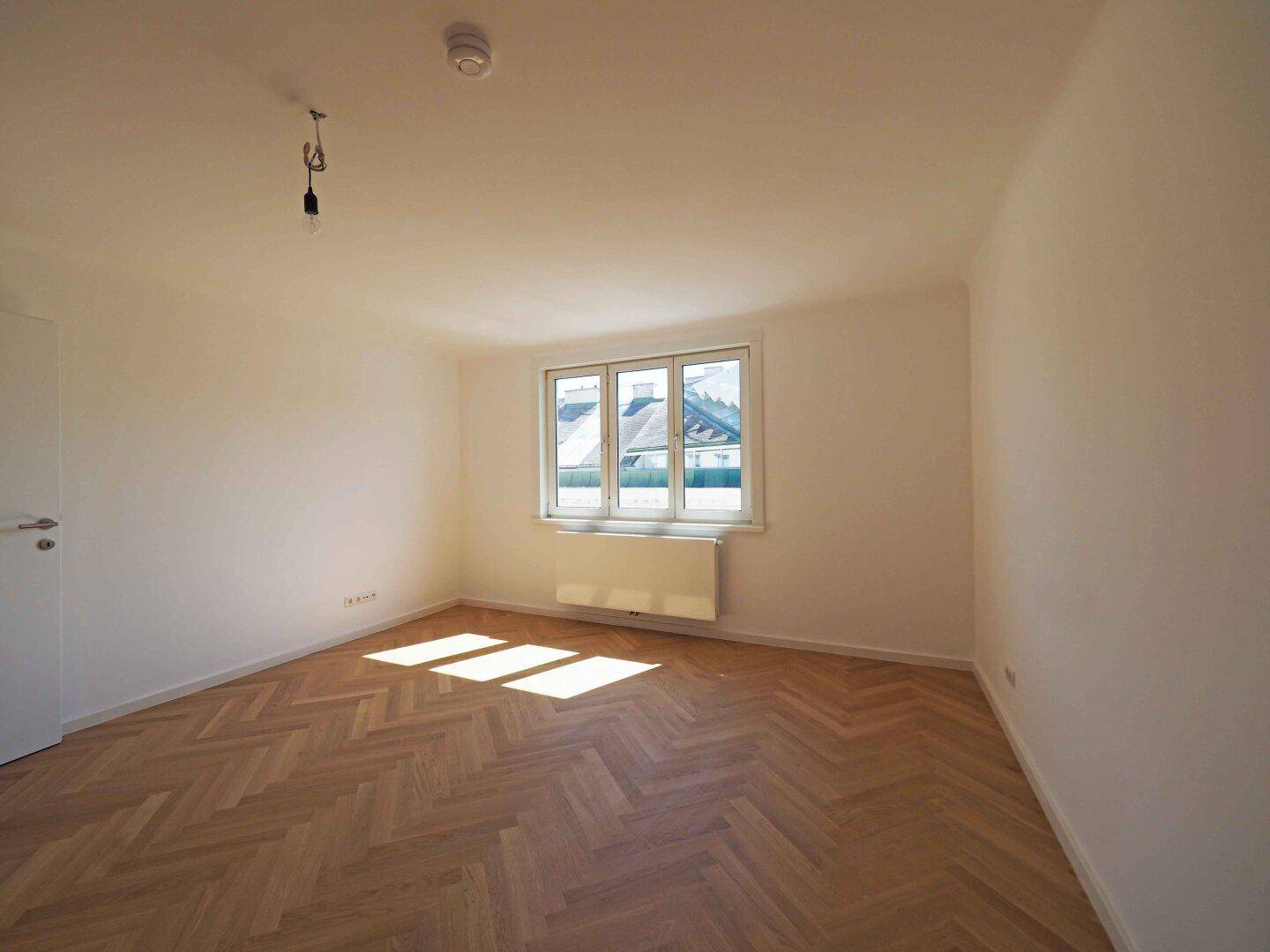 Helles Zimmer mit Fischgrätparkett und einem großen Fenster, bietet viel Tageslicht.