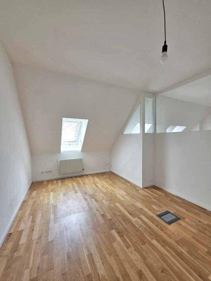 Helles Zimmer im Dachgeschoss mit schrägen Wänden, Holzparkettboden und einem Dachfenster.