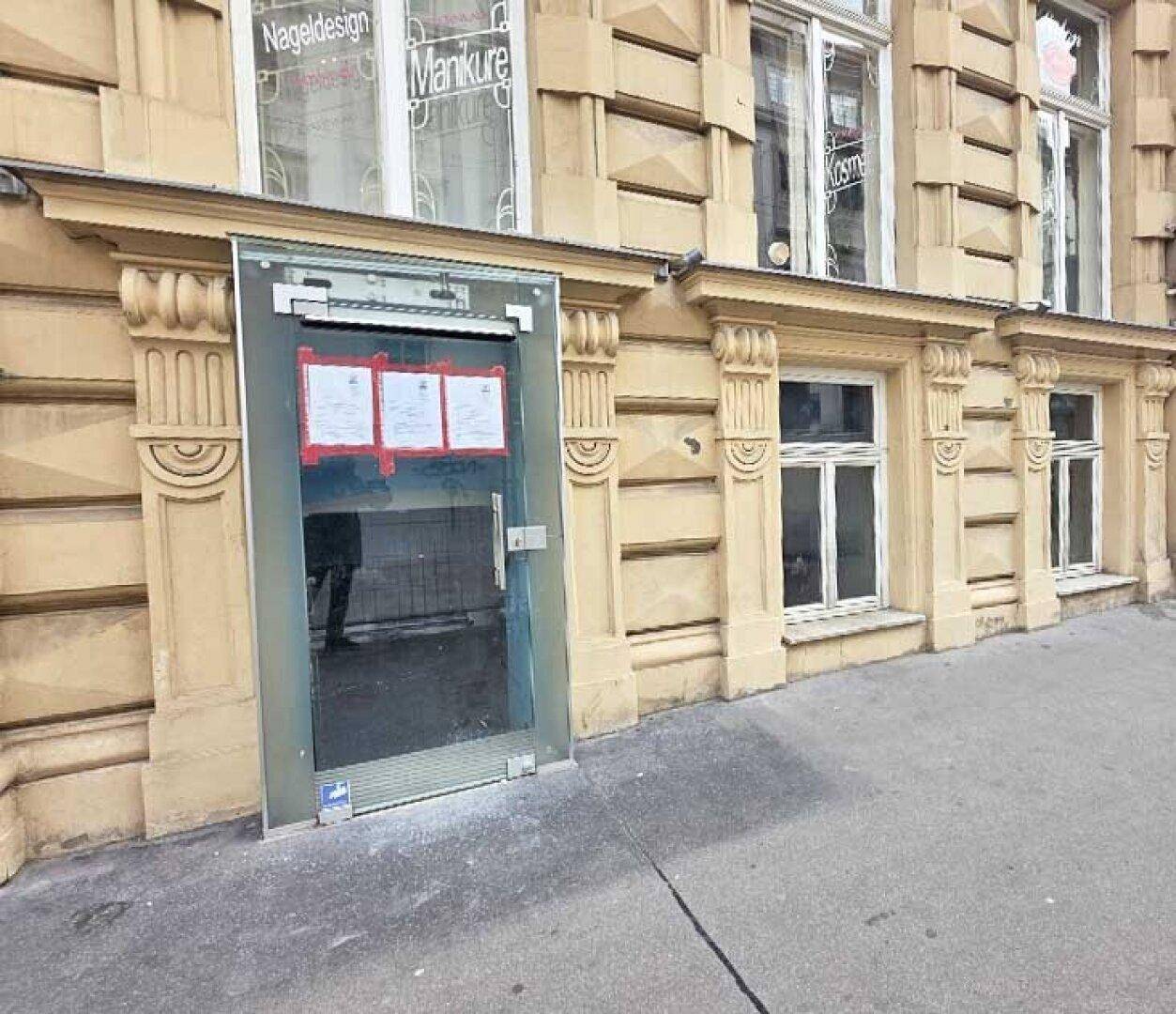 Nahaufnahme eines gläsernen Eingangsbereichs im Erdgeschoss mit Aushängen und der historischen Fassade.