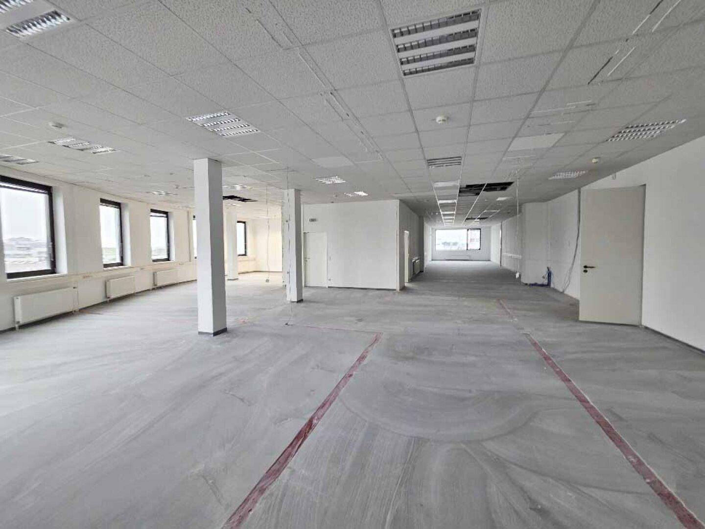 Geräumiges Büro mit Betonboden, abgehängter Decke und mehreren Fenstern, die viel Licht hereinlassen.