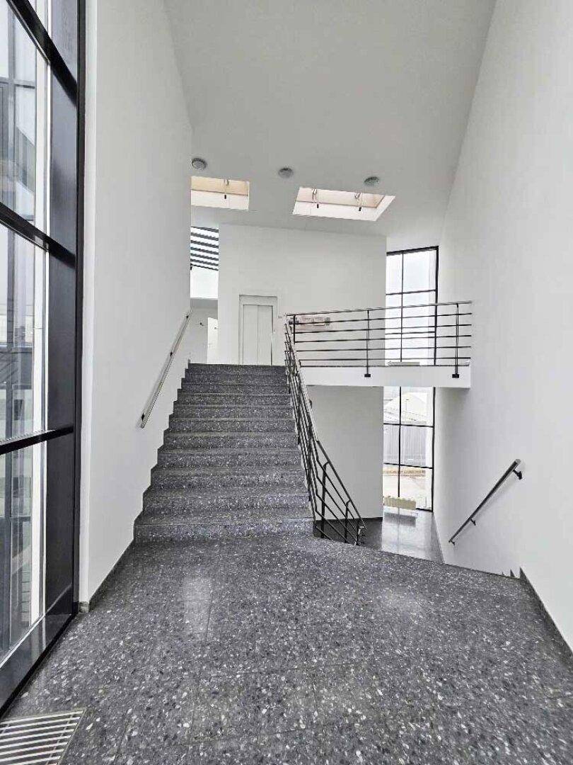 Moderner Treppenaufgang mit dunklem Terrazzoboden, großen Fenstern und einem Aufzug im Hintergrund.