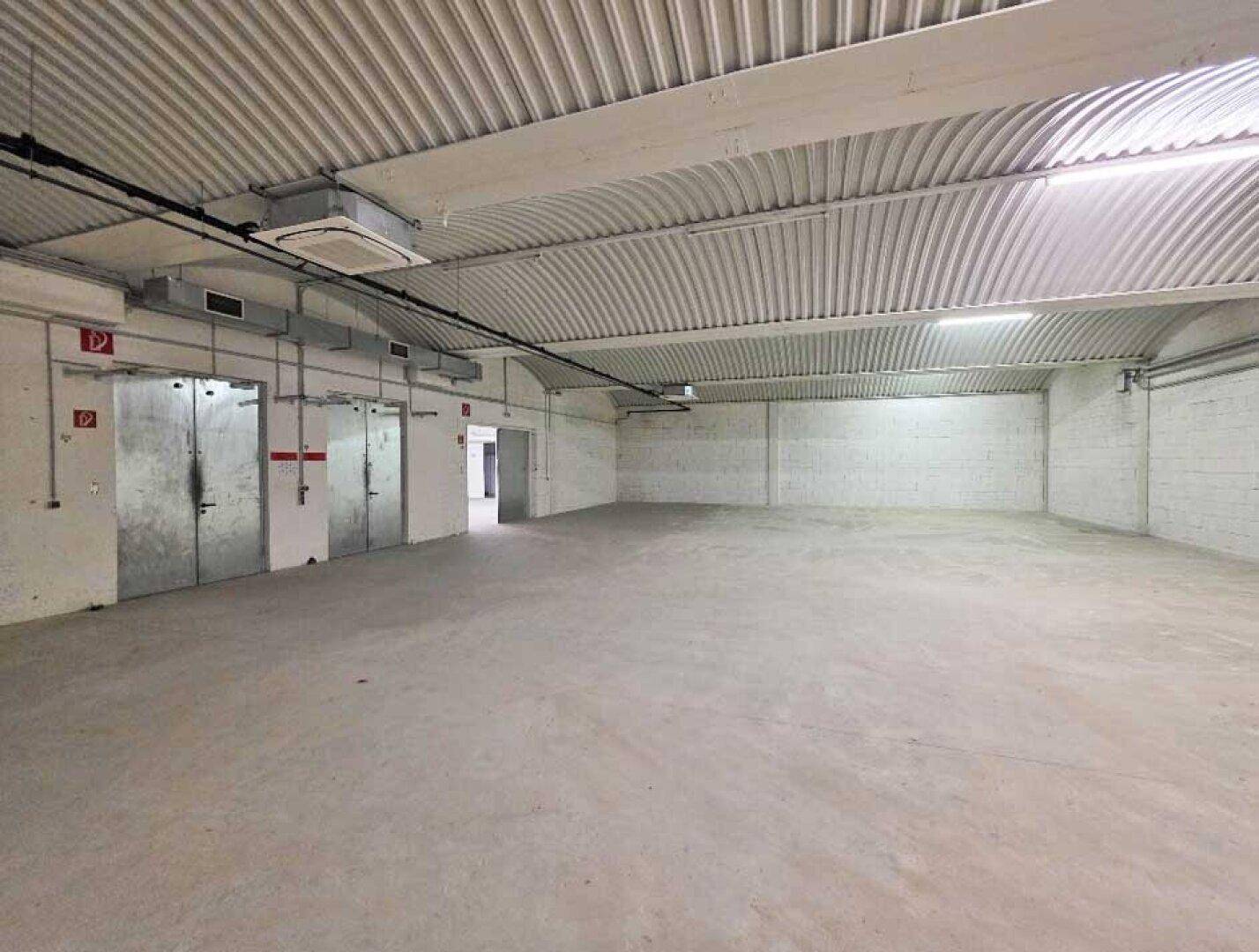 Große Lagerhalle mit gewölbter Decke, Betonboden und mehreren Türen im Hintergrund.