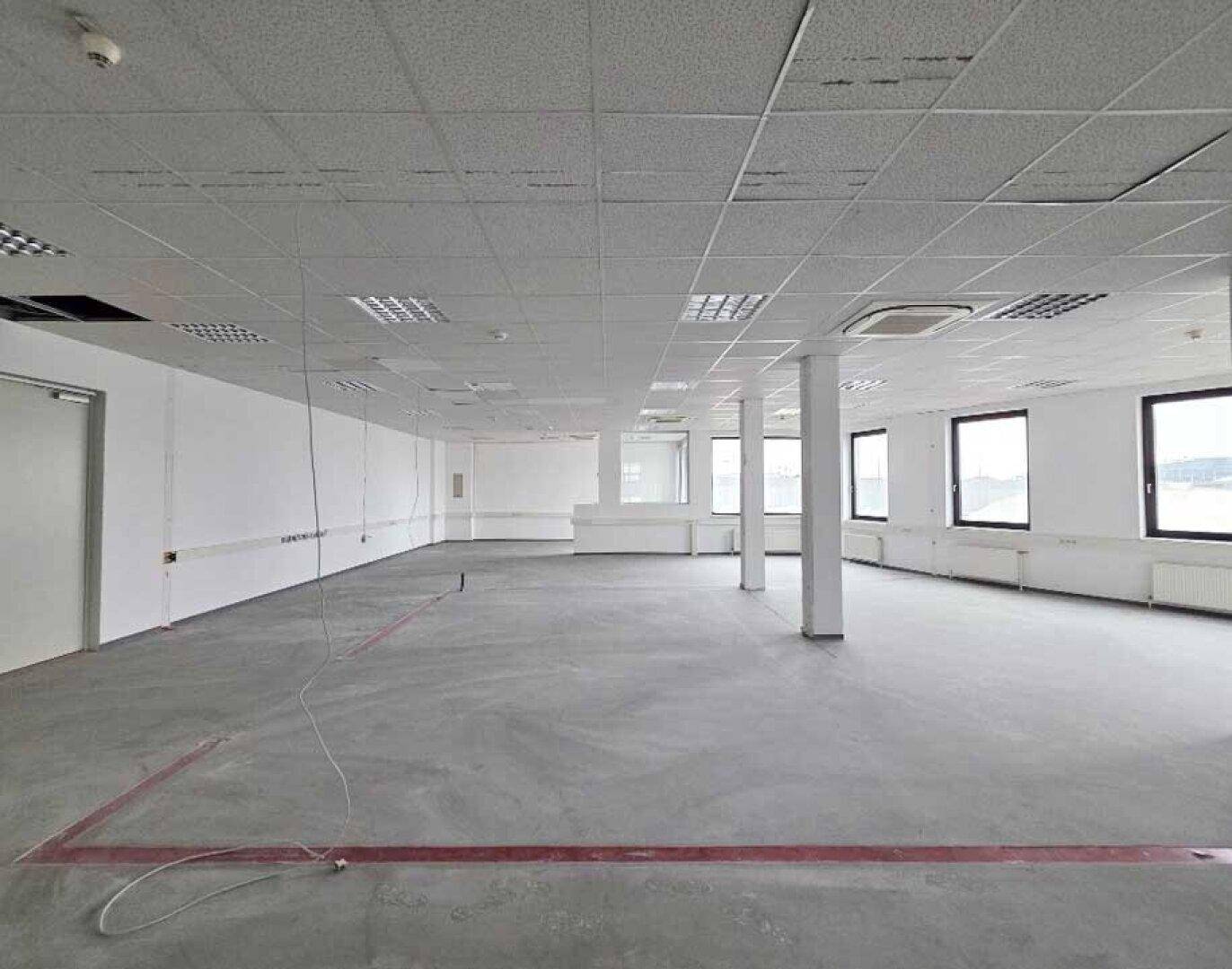 Helles, offenes Büro mit Säulen, Betonboden und einer Reihe von Fenstern entlang der Wand.