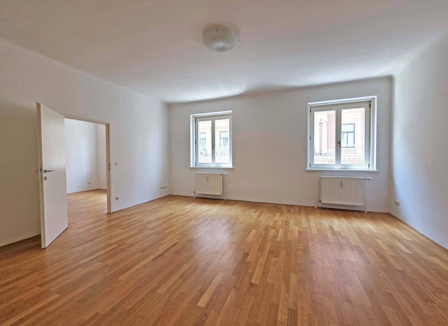 Geräumiges, leeres Wohnzimmer mit Parkettboden und zwei Fenstern.