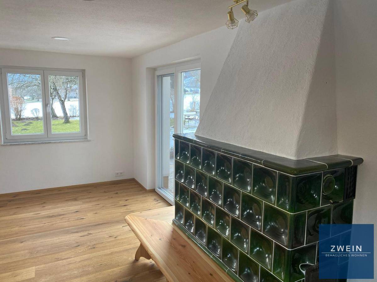 Gemütlicher Wohnraum mit traditionellem Kachelofen und Zugang zur Terrasse, hell und einladend.
