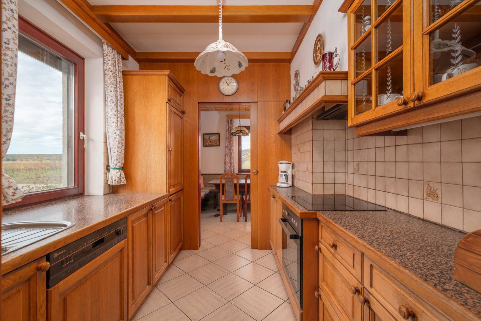 Küche mit Holzschränken, Fliesenboden, Einbaugeräten und Fenster mit Ausblick.