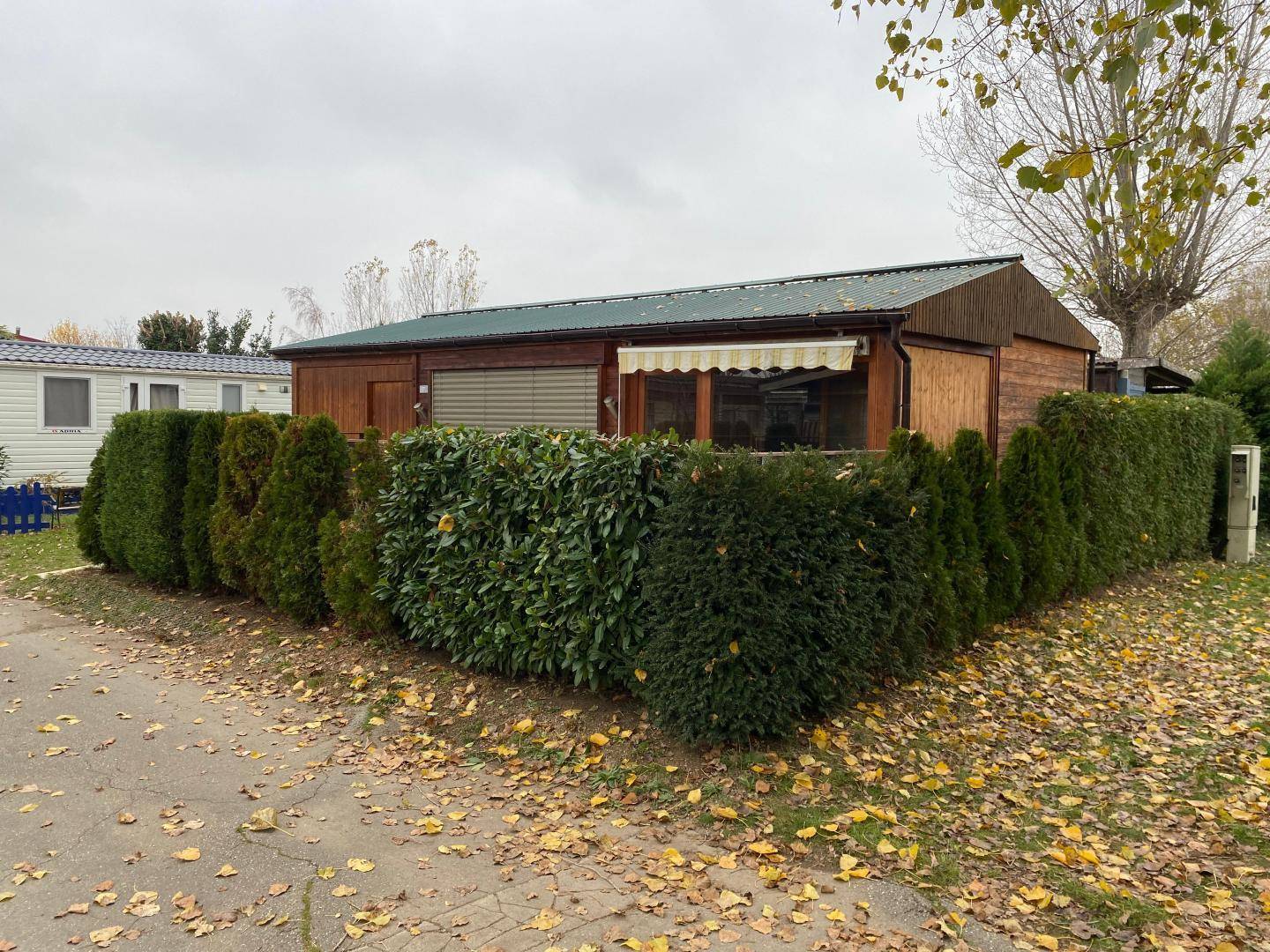 Holzhaus mit Markise und dichtem Heckenbewuchs, umgeben von Herbstlaub.