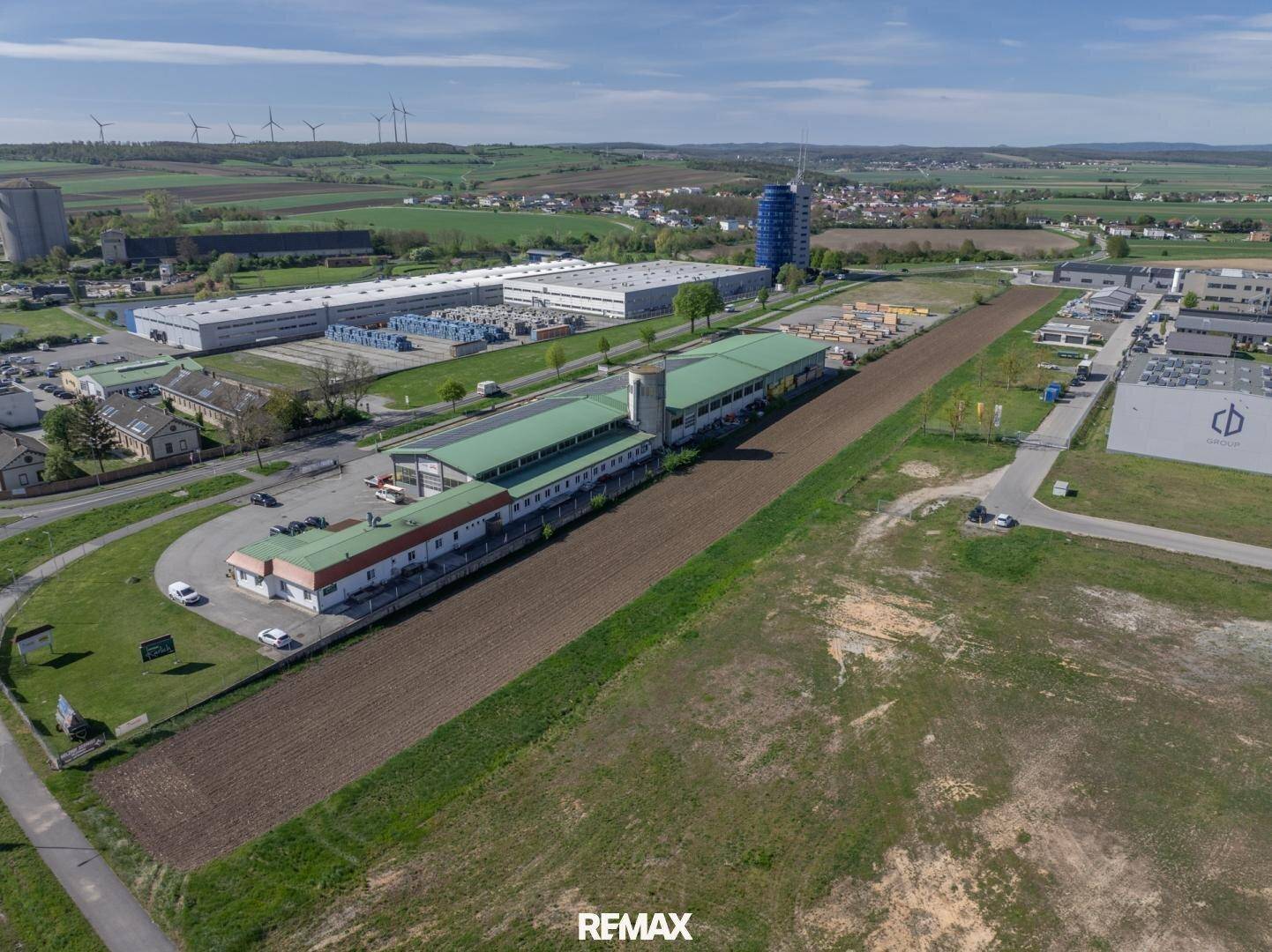 Weitläufige Luftaufnahme eines Gewerbegebiets mit Industriegebäuden und Windkraftanlagen im Hintergrund.