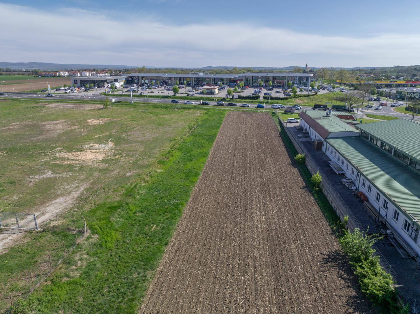 Nahaufnahme eines Ackerstreifens neben Gewerbegebäuden mit einem großen Parkplatz und Einkaufszentrum im Hintergrund.
