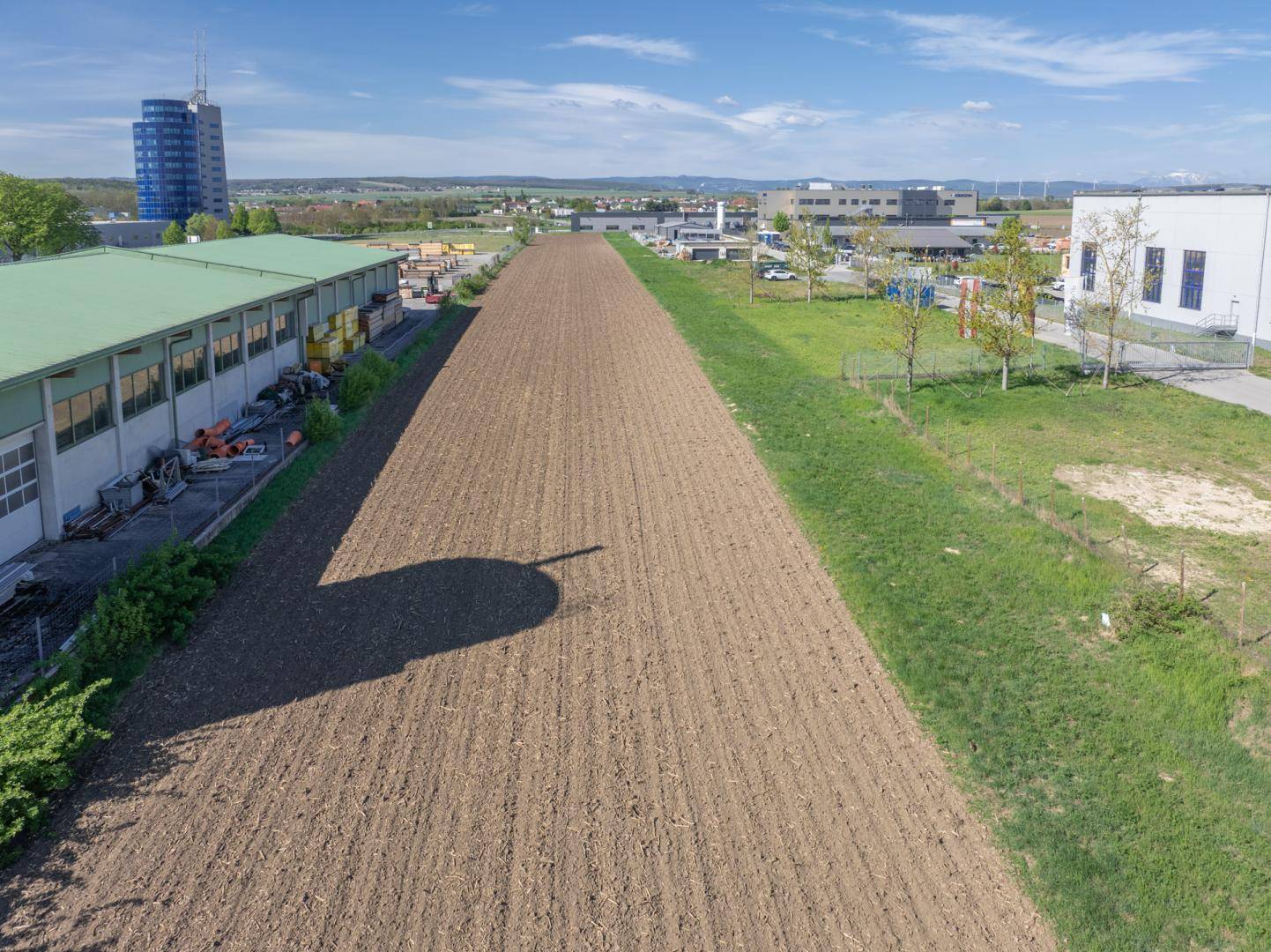 Detailansicht eines Ackerstreifens zwischen Gewerbegebäuden unter blauem Himmel mit einer Schattenprojektion.