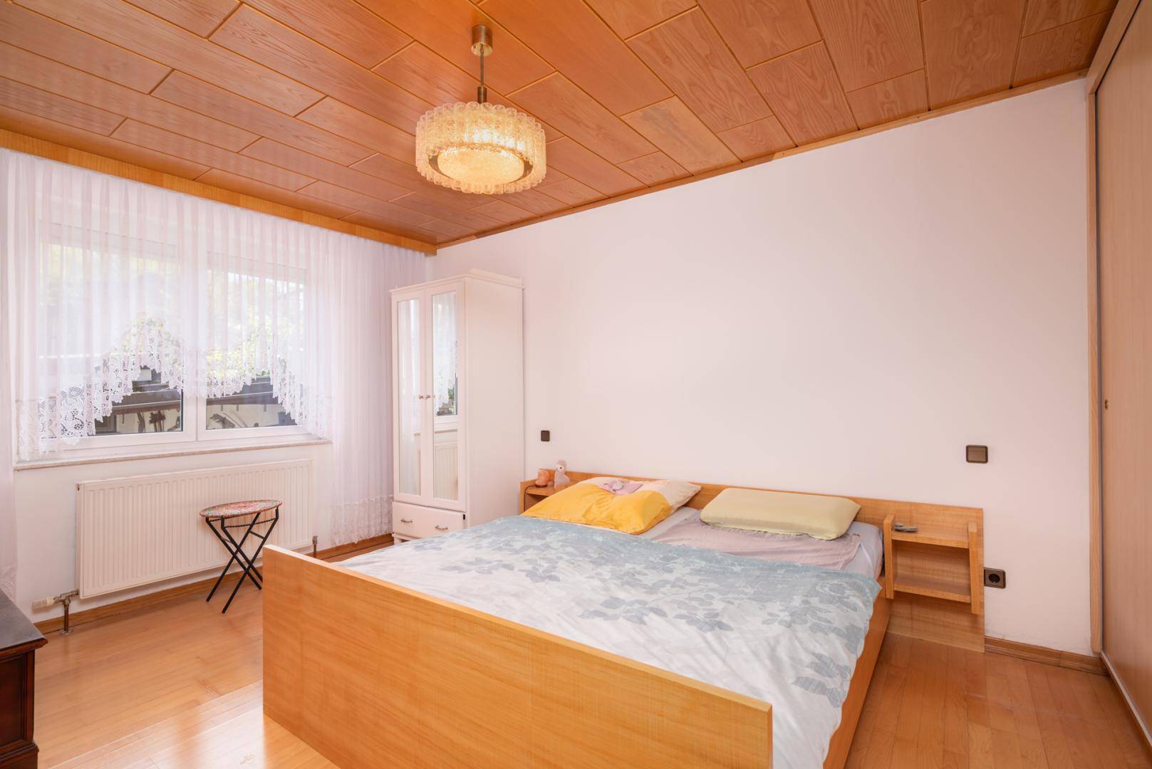 Helles Schlafzimmer mit einem Holzbett, weißem Kleiderschrank und einem Fenster, das viel Licht hereinlässt.