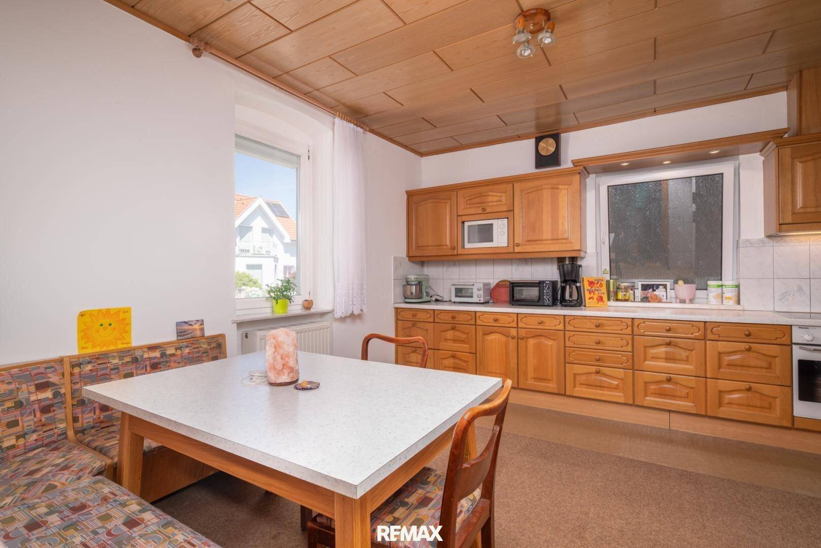 Traditionelle Wohnküche mit Holzschränken, Essbereich und Blick ins Grüne durch das Fenster.