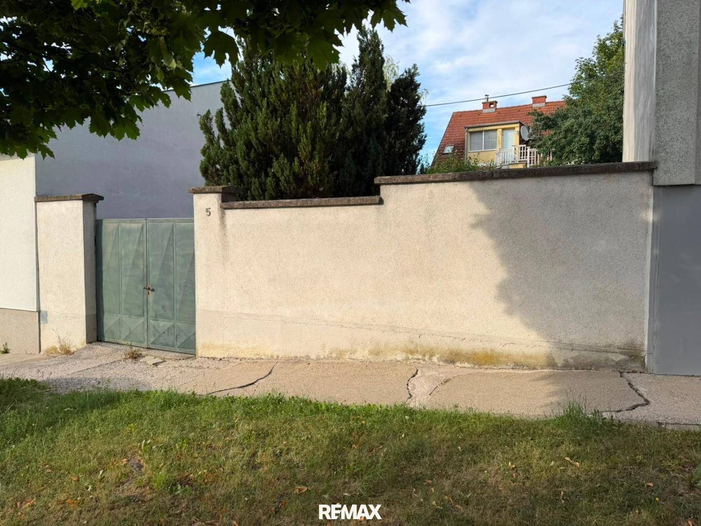 Eine hohe weiße Mauer mit einem grünen Metalltor und einem Haus im Hintergrund.