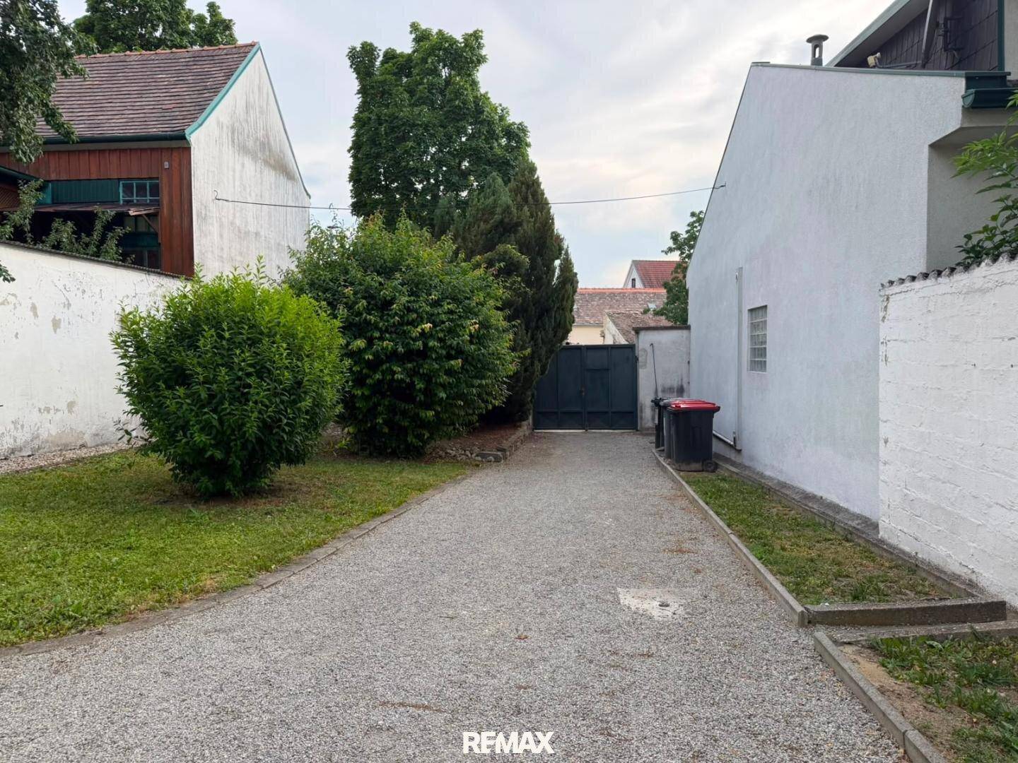 Kiesweg führt zu einem Tor, umgeben von grünen Büschen und einer weißen Mauer.