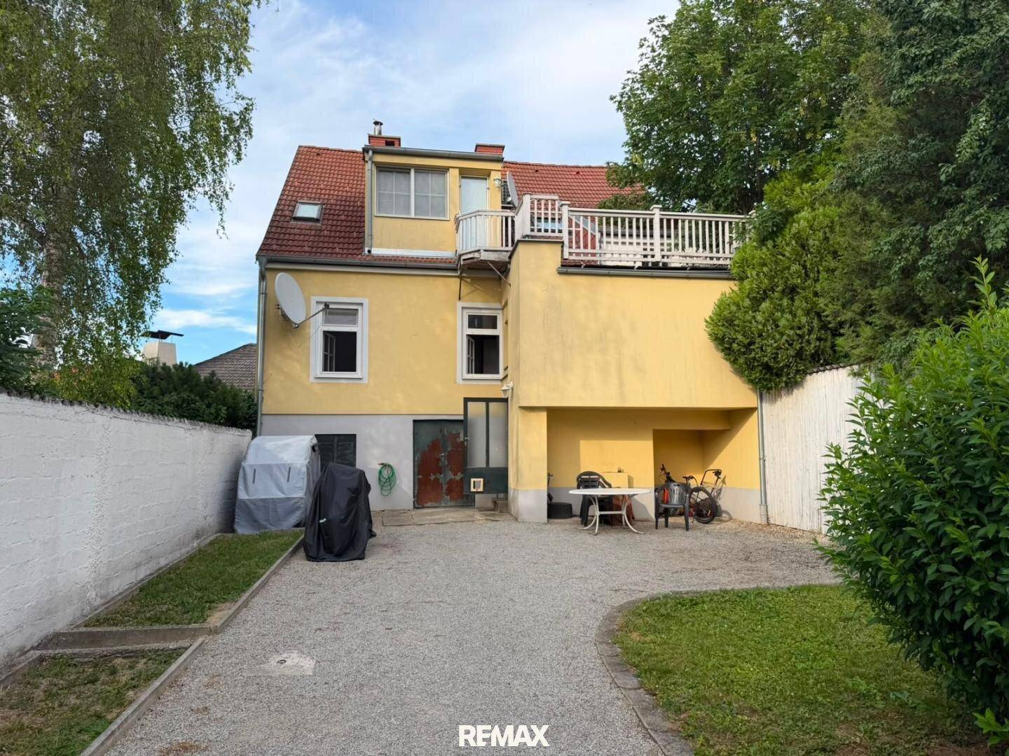 Hinterhof mit Kiesfläche, Rasen und einer Terrasse im Obergeschoss, umgeben von Bäumen.